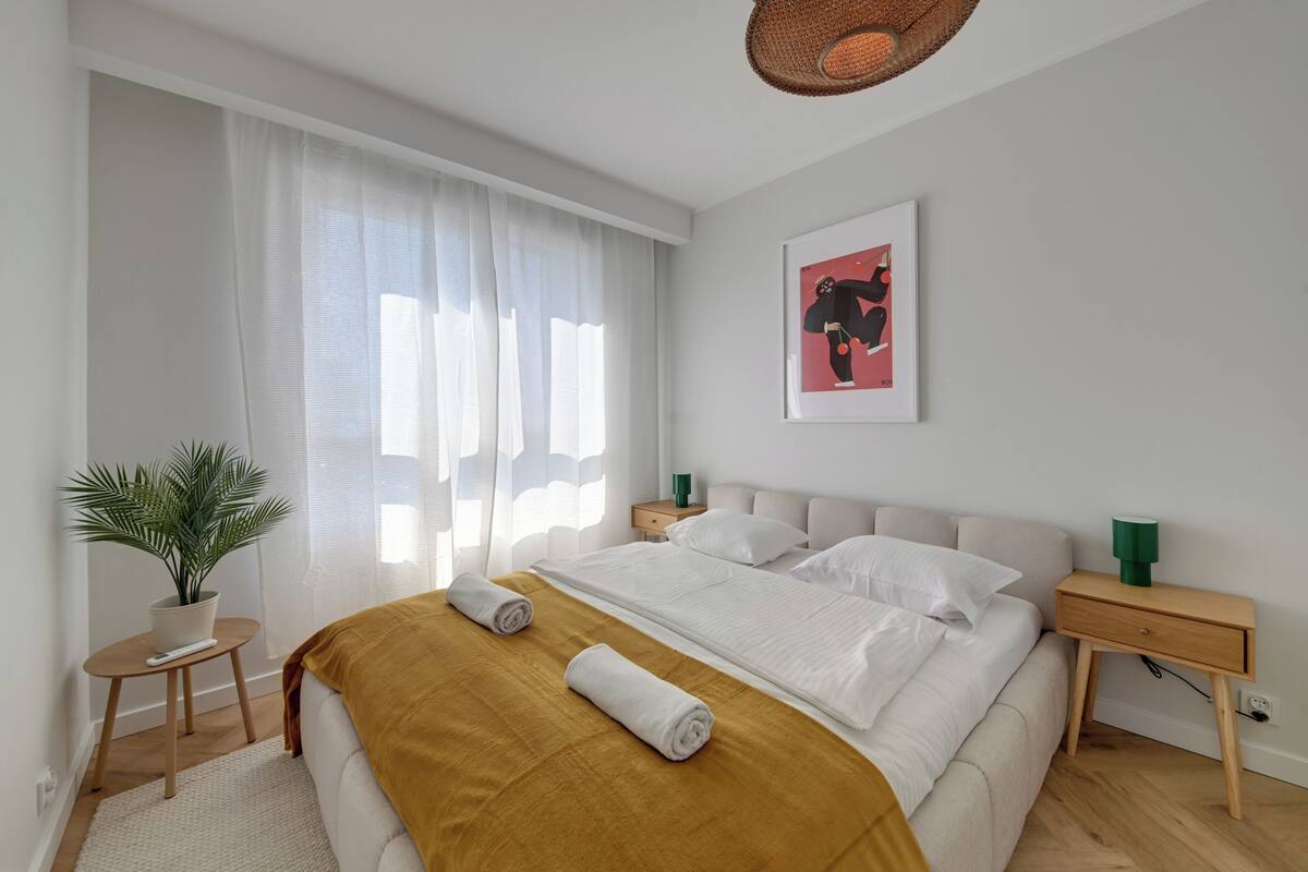 Une chambre moderne avec un grand lit, une table de chevet, une commode, une plante en pot et une fenêtre avec des rideaux légers.