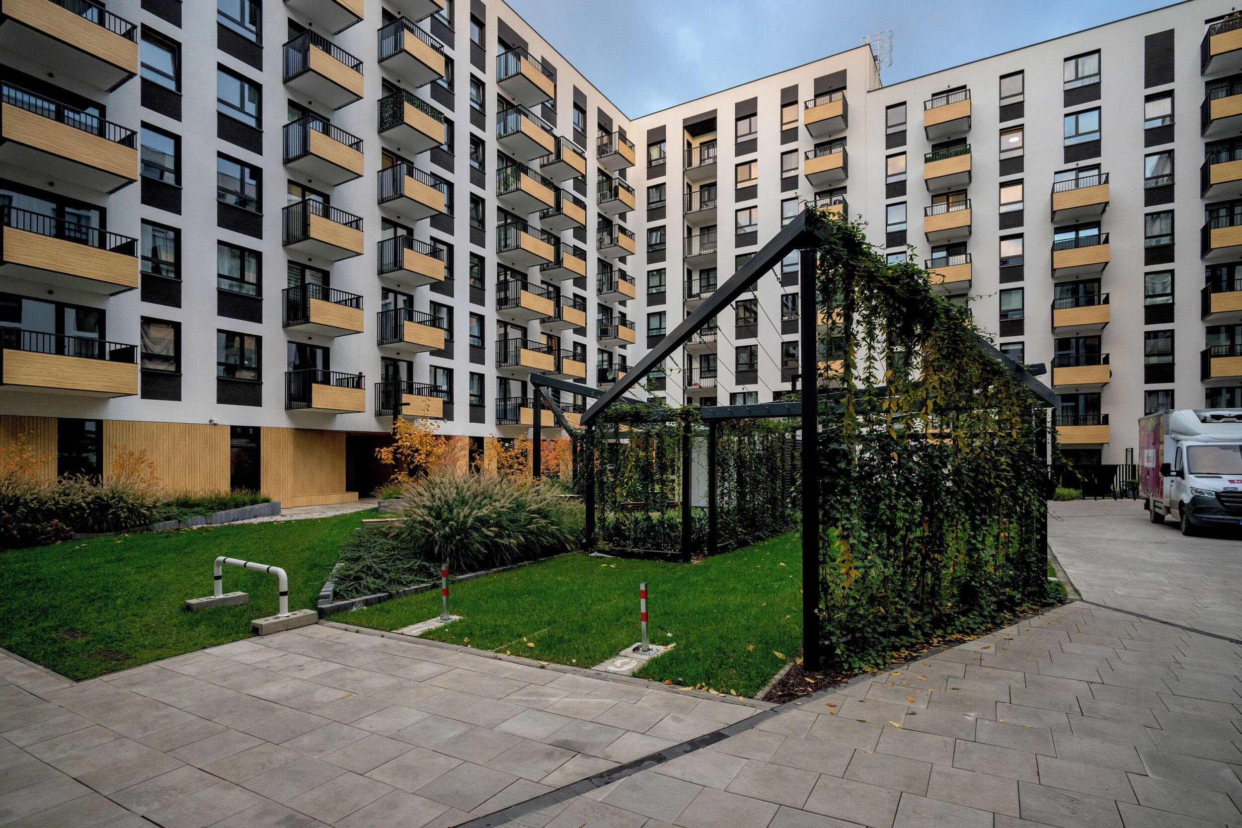A modern apartment building with multiple balconies, a small garden area with a pergola, and a paved walkway.