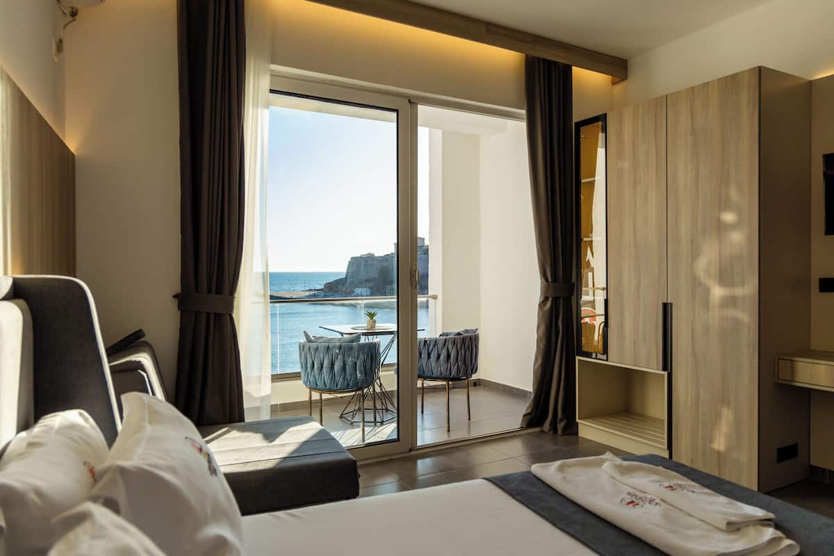 A hotel room with a bed, a small table with chairs, a view of the sea, and a wardrobe.