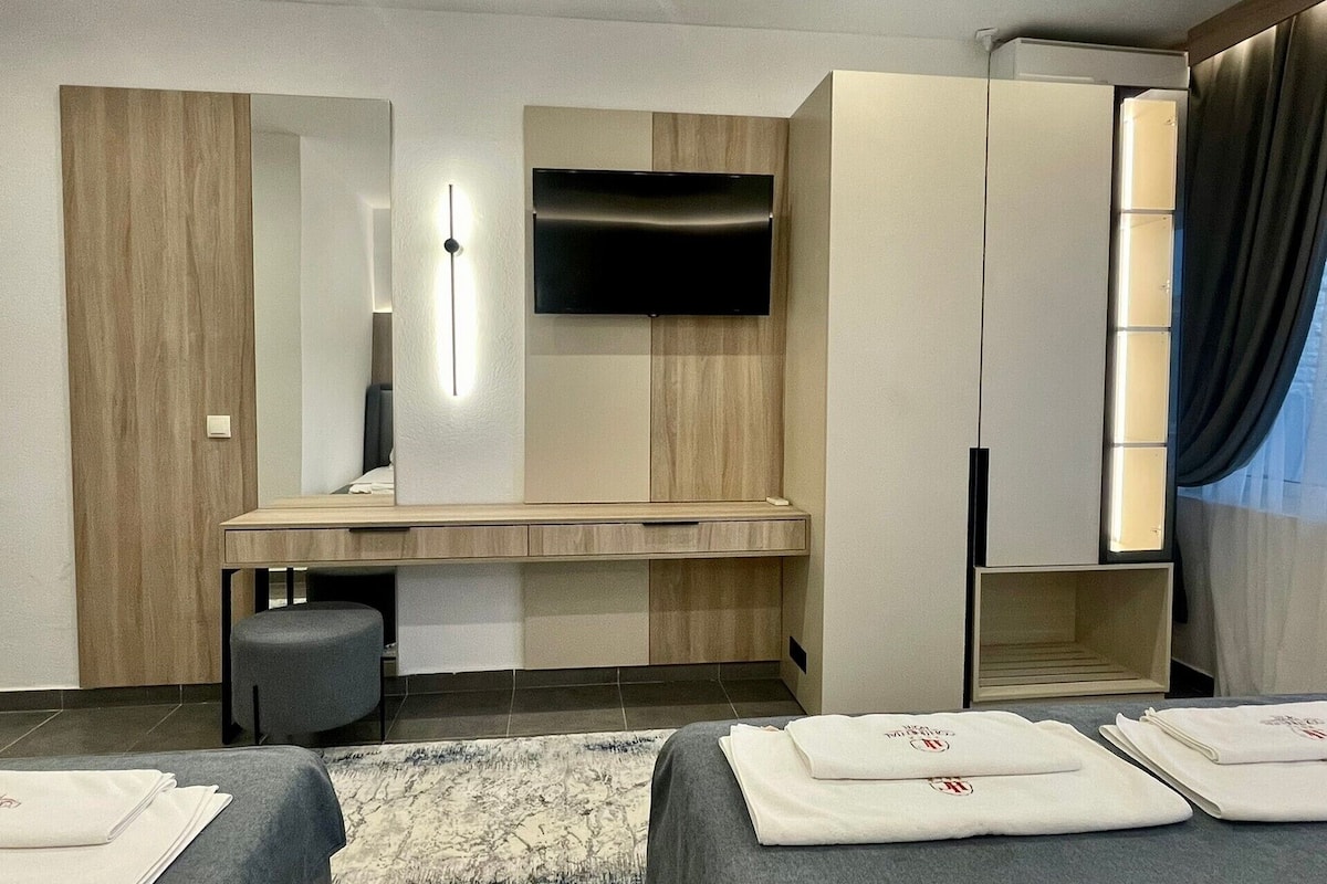 A modern hotel room with a flat-screen TV, a desk with drawers, a small stool, and neatly folded towels on the bed.