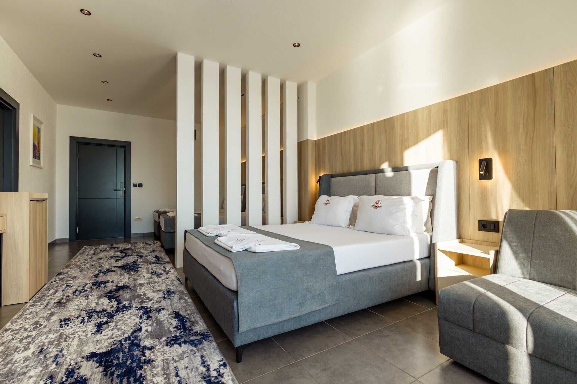 Una habitación de hotel moderna con una cama grande, un sofá gris, un tapete estampado y una pared de acento de madera.