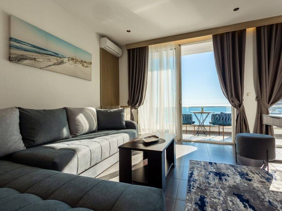 A modern living room with a grey sofa, a small black table, and a blue rug. There is a large window with curtains, a painting on the wall, and a view of the sea.