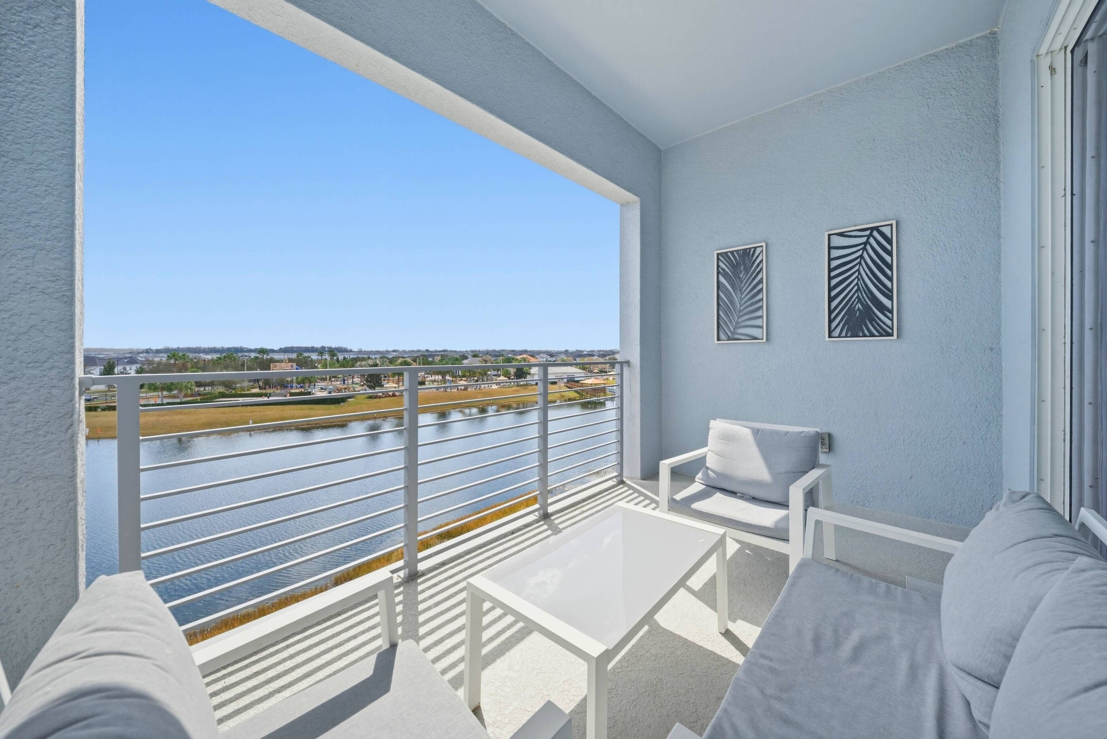 A balcony with a view of a river and distant buildings, white furniture, and two framed artworks.