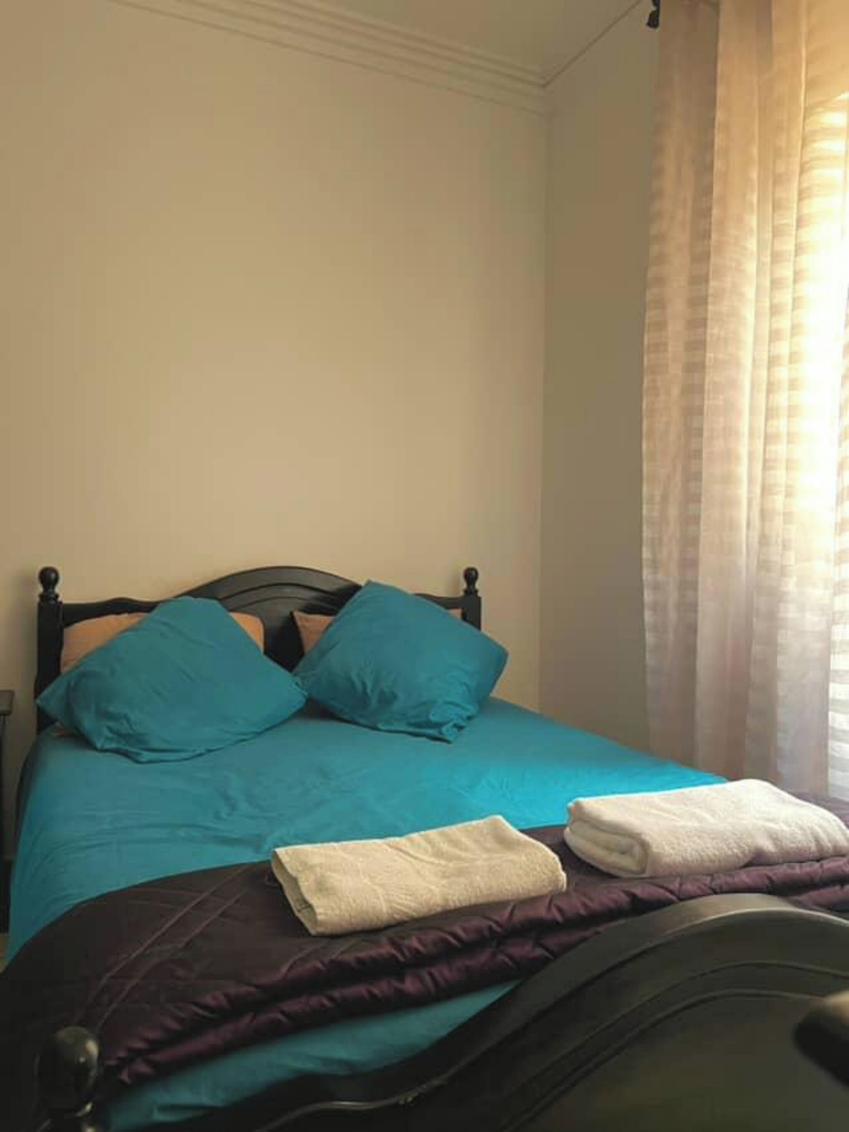 Un lit bien fait, avec une literie de couleur turquoise et deux oreillers, une serviette blanche pliée et une couverture violette posée sur une tête de lit de couleur foncée.