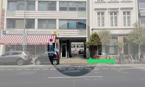 A street view with a building featuring a storefront with a red and white striped awning, a signboard with various business names, and a bicycle parking area.