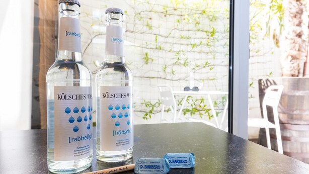Two bottles of water labeled 'KÖLSCHES WASS' and 'rabbelig' on a table with a wooden spoon and two chocolate bars.