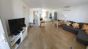 Un salon moderne avec un téléviseur à écran plat, un canapé agrémenté de coussins colorés, un coin repas avec une table et des chaises, et une cuisine en arrière-plan.