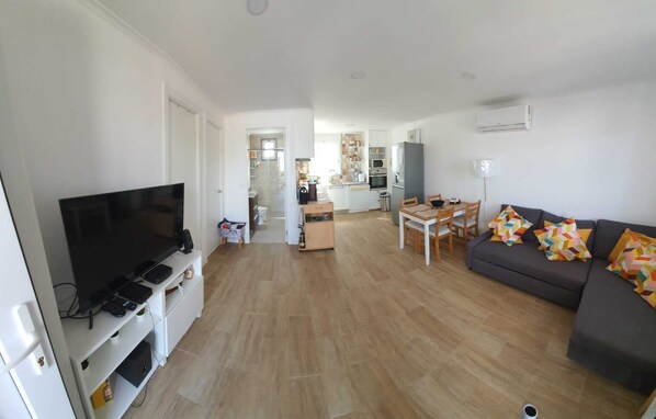 Una sala moderna con un televisor de pantalla plana, un sofá con cojines coloridos, una zona de comedor con mesa y sillas, y una cocina al fondo.