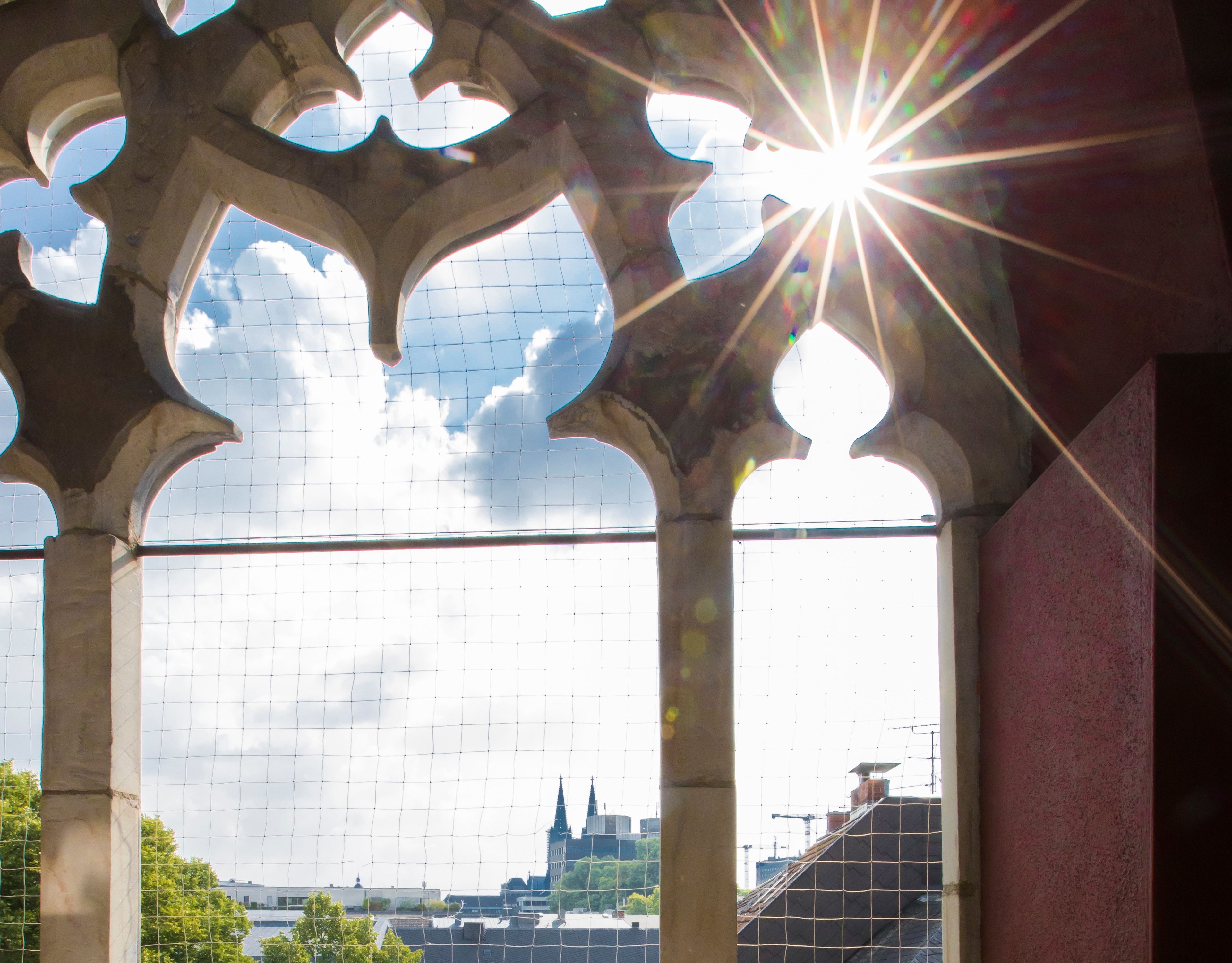 Ein gotisches Fenster mit Blick auf eine Stadtlandschaft, darunter ein Kirchturm.