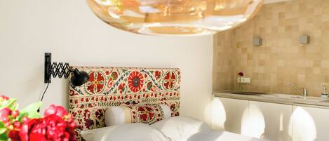 A hotel room with a bed, a decorative headboard, a small kitchenette, and a potted plant.