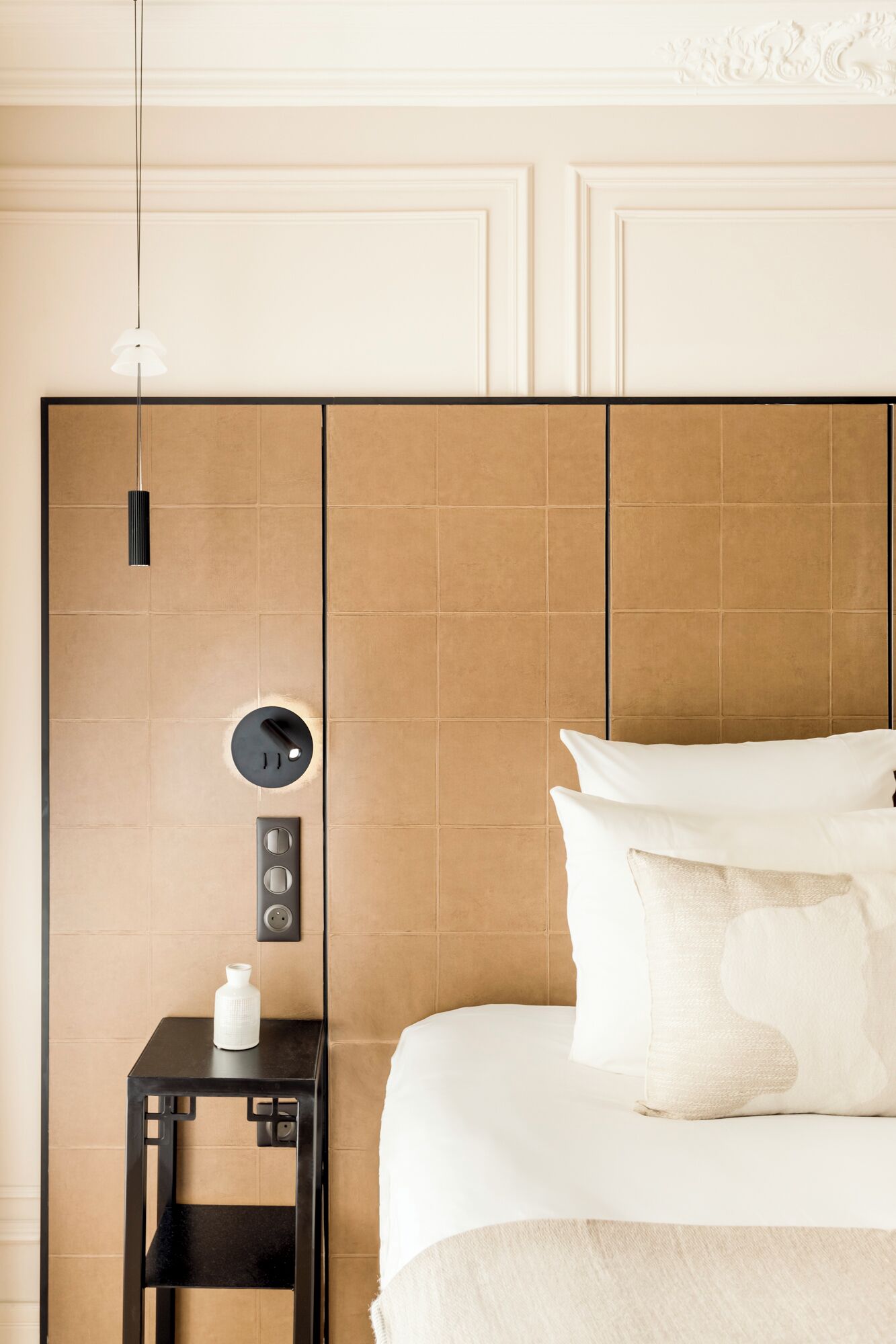 A modern hotel room with a large bed, a bedside table with a lamp, and a control panel on the wall.