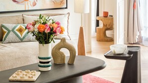 A modern living room with a large abstract painting, a round coffee table with decorative items, a sofa with patterned cushions, and a view into the adjacent room. - Merveil Luxury Suite - Bvd Saint Denis (Paris)