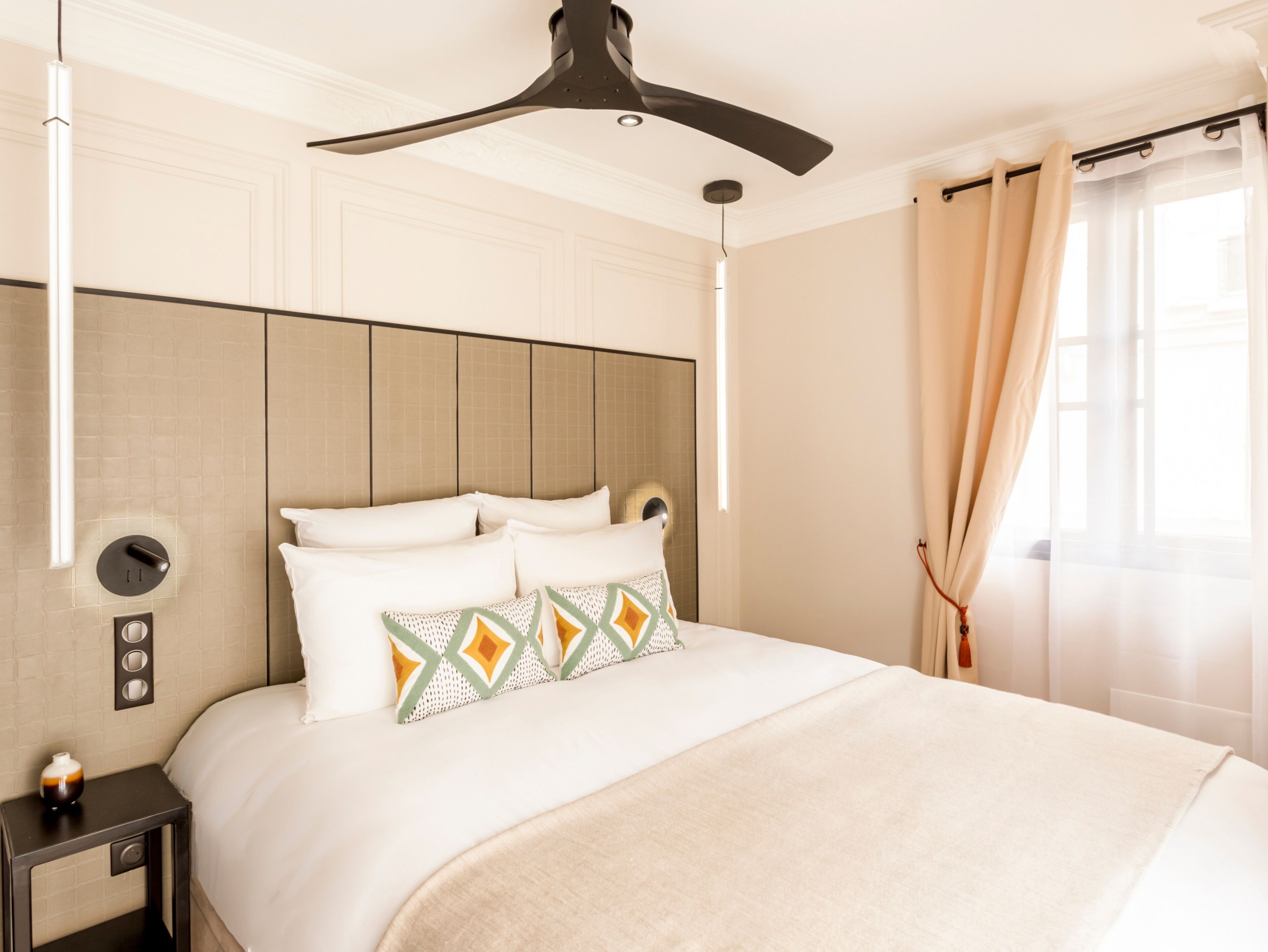 A hotel room with a large bed, a ceiling fan, a bedside table with a vase, and a window with curtains.