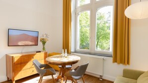 A room with a round wooden table, two grey chairs, a wooden cabinet, a television, and a window with curtains.