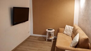 Un salon avec un téléviseur à écran plat fixé au mur, une petite table d’appoint blanche avec une télécommande, et un canapé beige agrémenté de coussins.