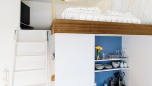 A kitchen with white cabinets, a blue wall, and a wooden ceiling. There are shelves with dishes, glasses, and utensils. - my.statthaus - 8 apartments exclusively for up to 22 people (Köln)