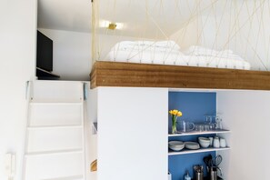 Une cuisine avec des meubles blancs, un mur bleu et un plafond en bois. On y trouve des étagères remplies d’assiettes, de verres et d’ustensiles de cuisine.
