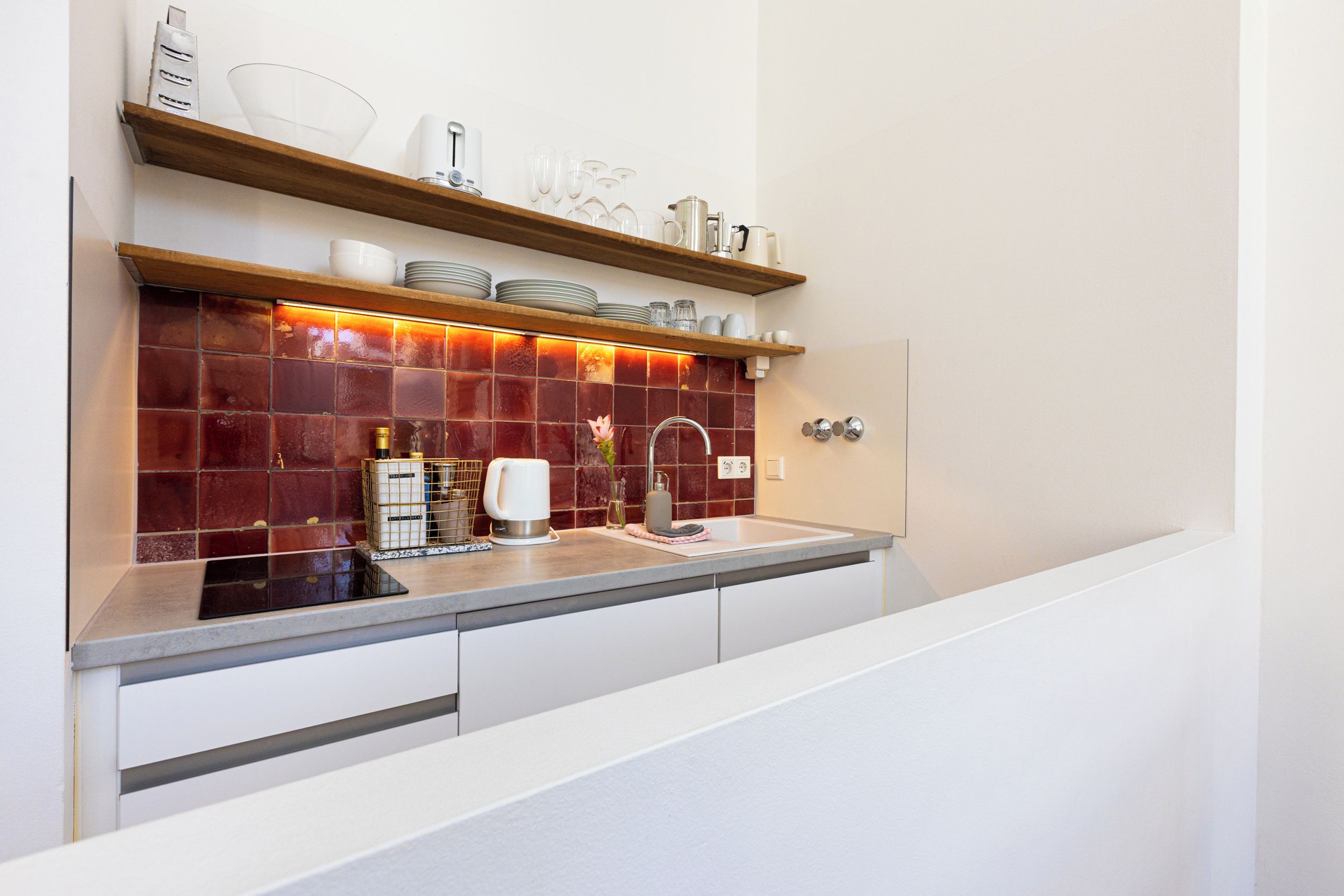 A modern kitchen with a red-tiled backsplash, stainless steel appliances, and wooden shelves above the counter.