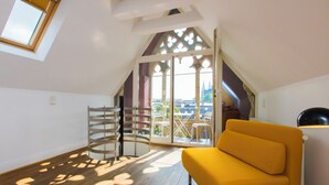 Une pièce avec un canapé jaune, un plancher en bois, une porte en métal et une vue sur la ville par une grande fenêtre.