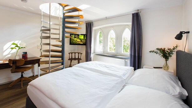 Une chambre moderne avec un escalier en colimaçon, un lit avec du linge de maison blanc, un bureau avec une chaise, une télévision et une fenêtre avec des rideaux.