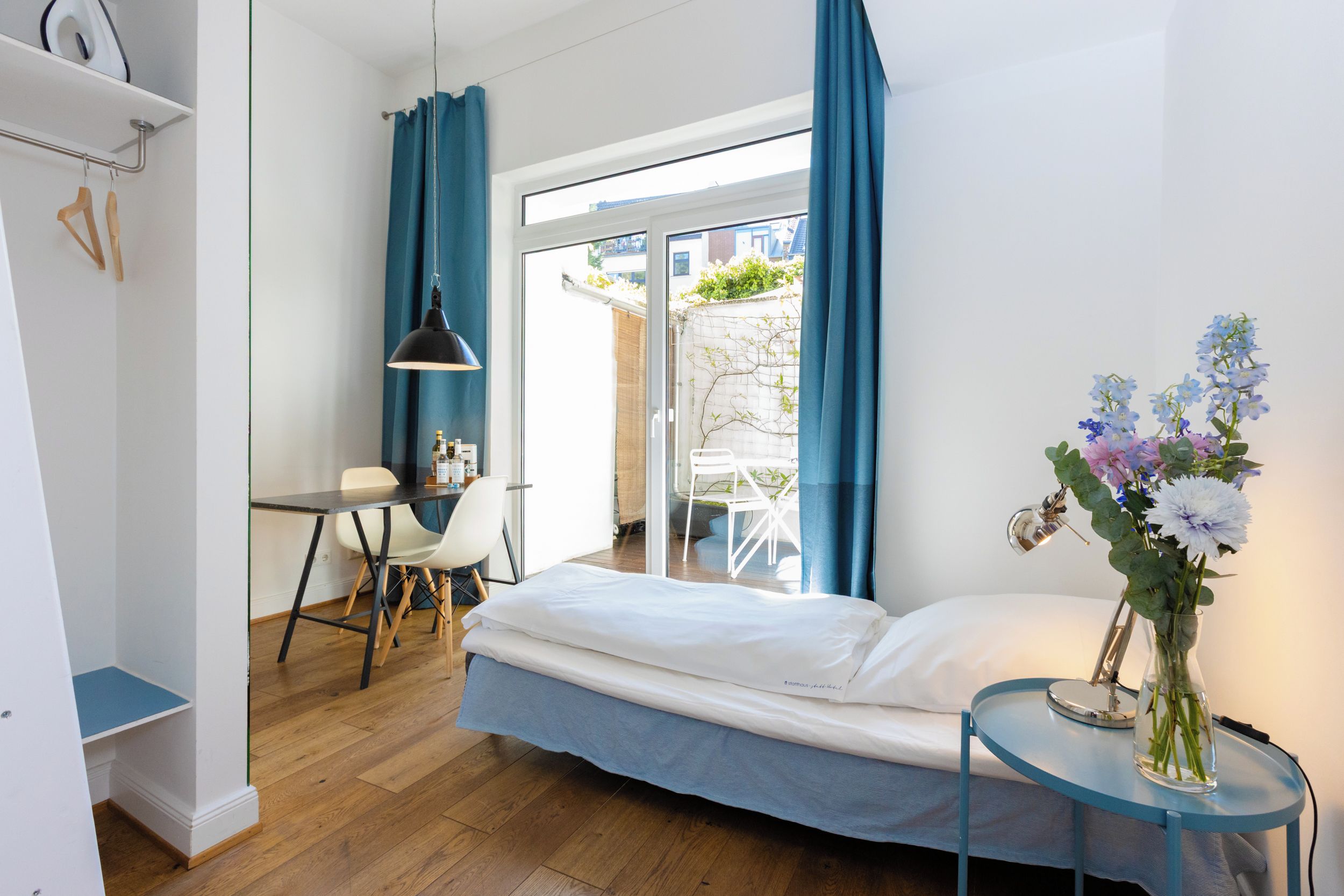 A bedroom with a bed, a desk, chairs, a hanging lamp, and a vase with flowers.