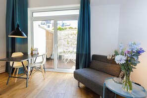 A modern living room with a sofa, a dining table with chairs, a pendant light, and a view through a glass door to an outdoor area. - my.statthaus - 8 apartments exclusively for up to 22 people (Köln)