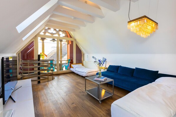 A modern loft bedroom with a sloped ceiling, a large window, a blue sofa, a small table, and a chandelier.