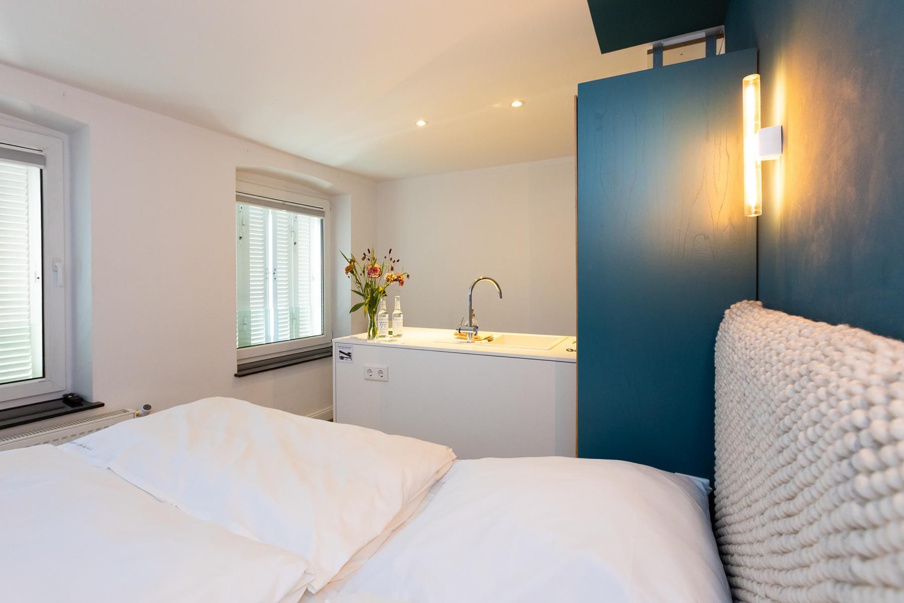 Une chambre d’hôtel comprenant un lit, une fenêtre avec des stores, un lavabo avec un miroir et un luminaire fixé au mur.