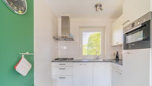 A modern kitchen with white cabinets, a stainless steel range hood, a Bosch oven, and a window with blinds. - Tastefully Furnished Detached Bungalow (De Koog)