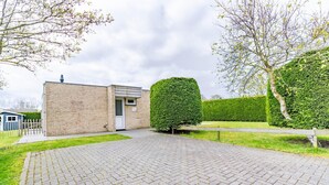 A paved driveway leading to a brick building with a small door, surrounded by well-maintained hedges and a tree. - Tastefully Furnished Detached Bungalow (De Koog)