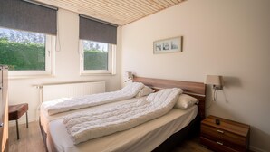A bedroom with a double bed, a bedside table, a nightstand, a lamp, and a window with a view of greenery. - Tastefully Furnished Detached Bungalow (De Koog)