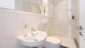 A modern bathroom with a white sink, toilet, and glass-enclosed shower. - Small studio apartment with a view over Cologne (Köln)