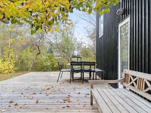 Eine Holzterrasse mit Tisch und Stühlen, umgeben von Bäumen und einem schwarzen Gebäude.
