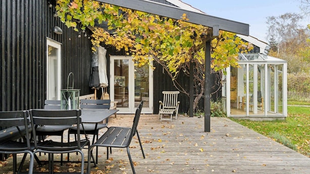 Een modern huis met een houten terras, een glazen kas en een pergola begroeid met wijnranken.