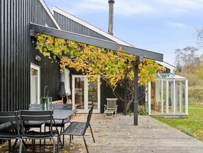 Ein modernes Haus mit einer Holzterrasse, einem Glashaus und einer mit Wein bewachsenen Pergola.