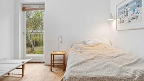 Een minimalistische slaapkamer met een bed, een klein tafeltje, een lamp en een raam met uitzicht op bomen.