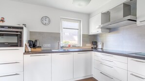 A modern kitchen with white cabinets, a built-in oven, a window with blinds, and a clock on the wall. - Charming Villa Near the sea and Forest (De Koog)