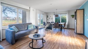 A spacious living area with a sofa, coffee table, and dining table. Large windows provide natural light and a view of the outdoors. - Charming Villa Near the sea and Forest (De Koog)