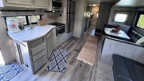 Cocina compacta para autocaravana con kitchenette, horno empotrado, microondas y refrigerador. Hay una ventana que proporciona luz natural y una pequeña zona de estar con mesa y cojines.