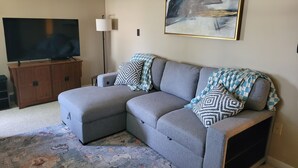 Un soggiorno con un divano angolare grigio, una televisione a schermo piatto su un mobile in legno, un tappeto decorato e un grande quadro astratto appeso al muro.