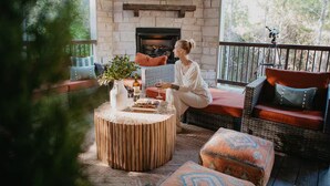 A cozy outdoor seating area with a stone fireplace, wicker furniture, and a television mounted above the fireplace. - Movie under Stars, Heated Pool, Pickle ball, Slide (Broken Bow)