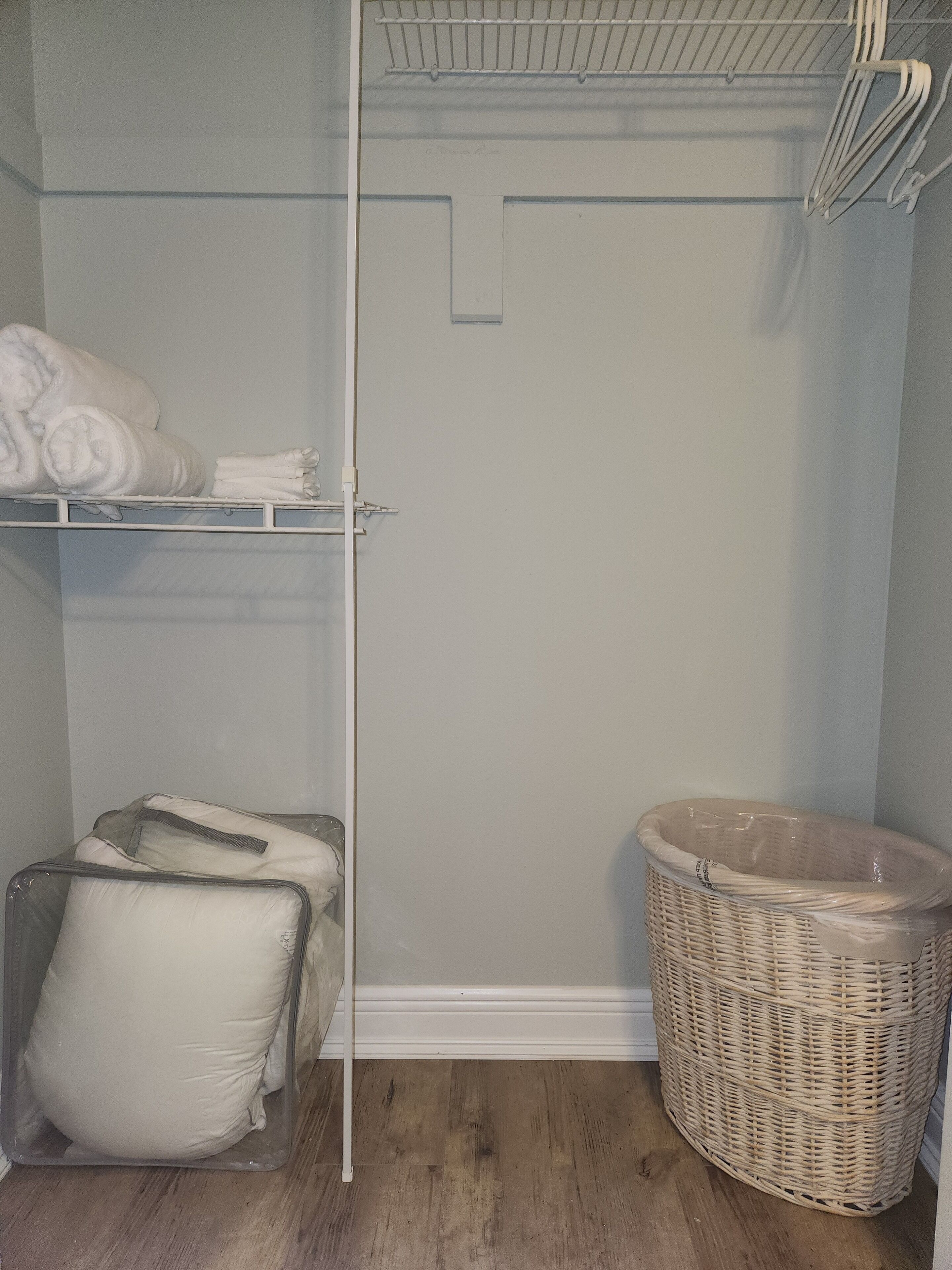 A small laundry room with a white shelf holding folded towels, a wicker laundry basket, and a clear plastic chair.