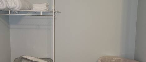 A small laundry room with a white shelf holding folded towels, a wicker laundry basket, and a clear plastic chair.