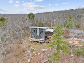 A modern two-story house with a deck, surrounded by a forested area.