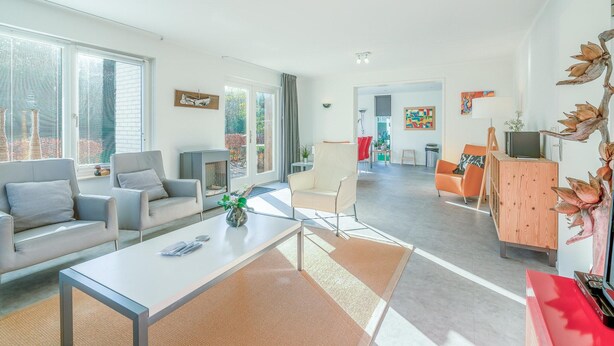 Een moderne woonkamer met een bank, fauteuils, een salontafel en een televisie. Achtergrond is er een open haard, een eethoek en een keuken.