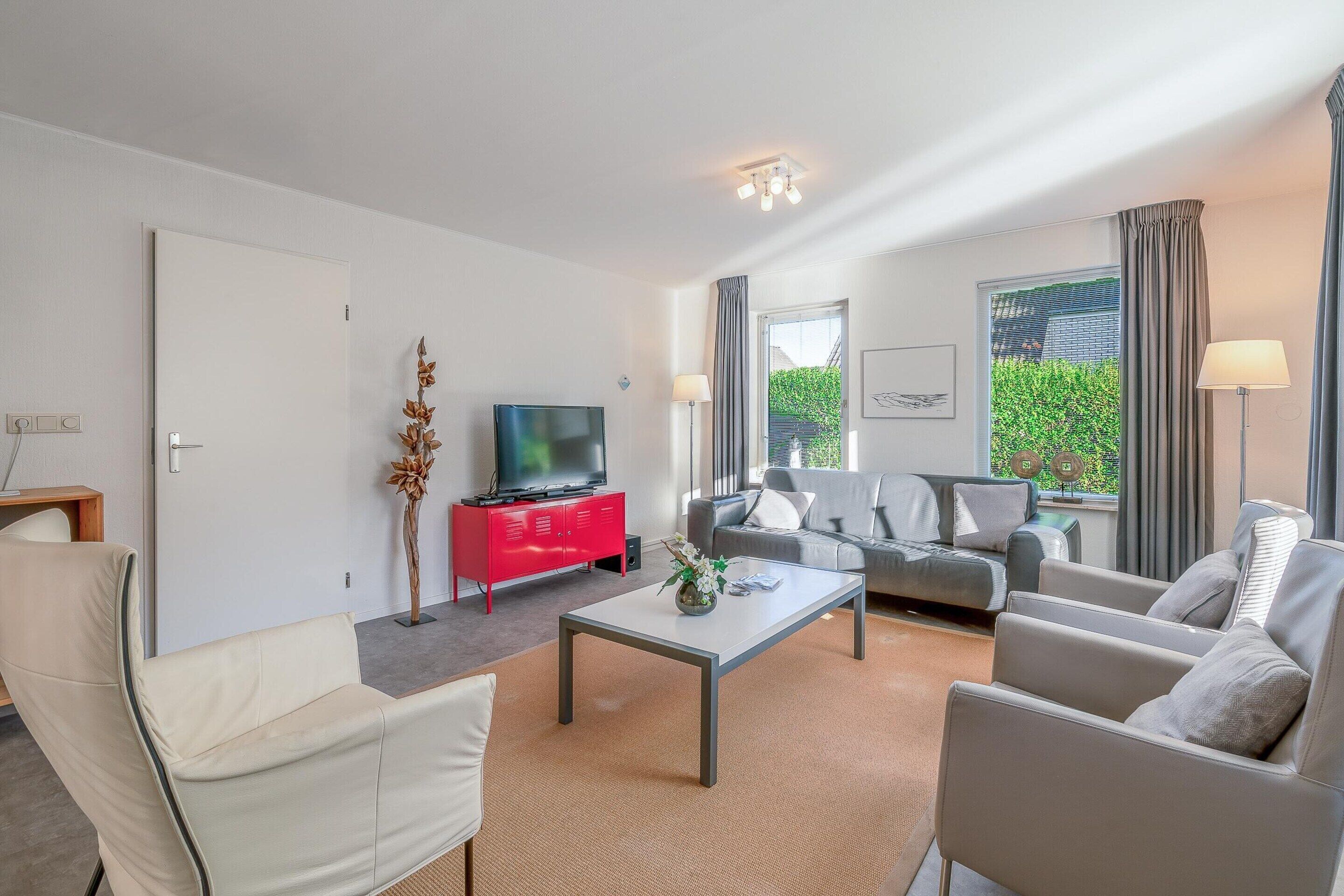 A modern living room with a sofa, armchairs, a coffee table, a red cabinet, a flat-screen TV, and a large window with curtains.
