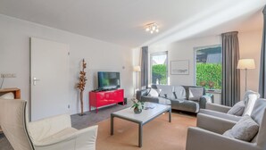 Een moderne woonkamer met een bank, fauteuils, een salontafel, een rode kast, een flatscreen televisie en een groot raam met gordijnen.