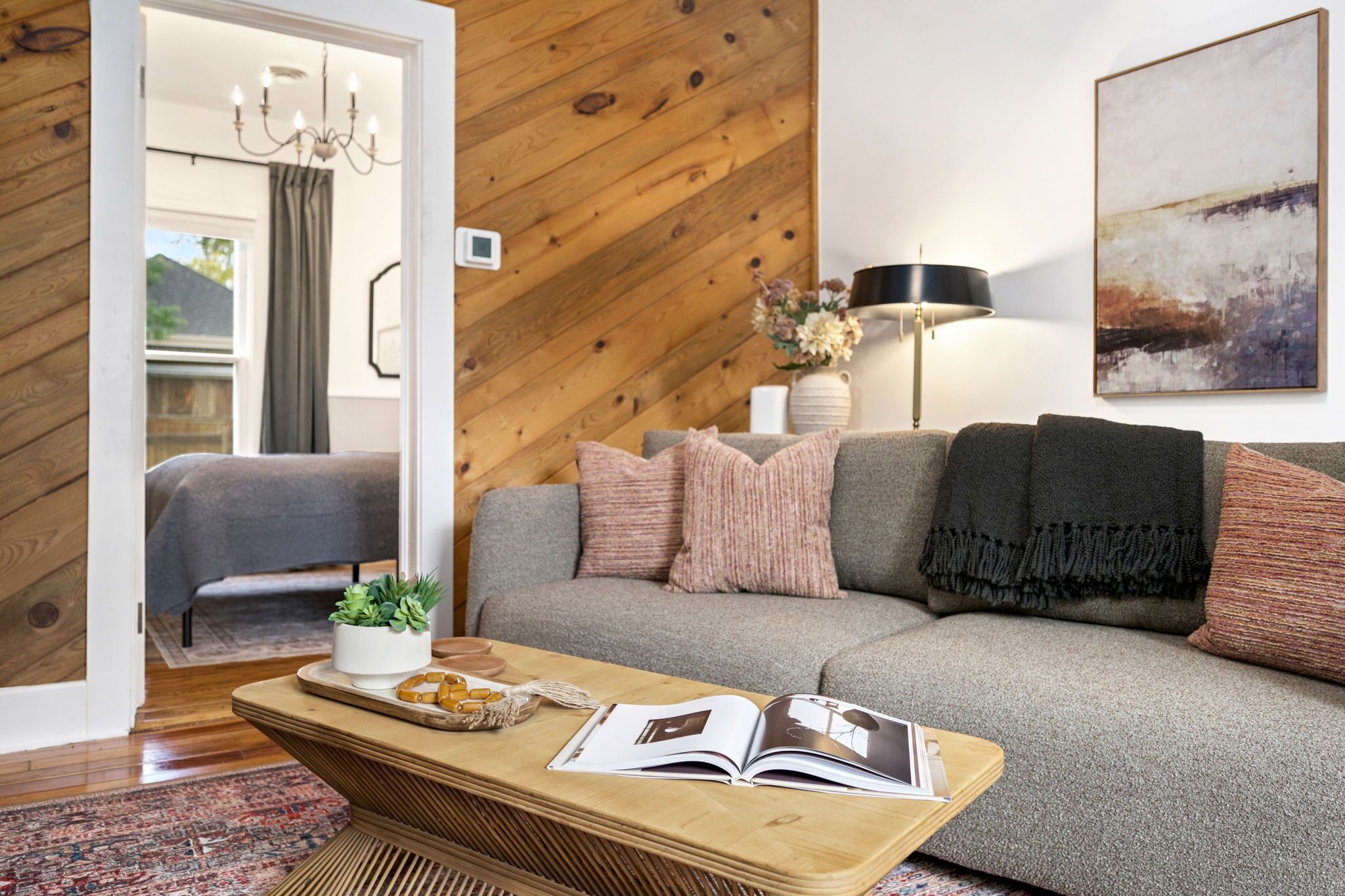 Un salón acogedor con un sofá gris, una mesa de centro de madera con un libro y objetos decorativos, una lámpara y un cuadro en la pared.