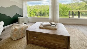Quarto com uma janela grande, cômoda de madeira, um pufe estampado e vista para o verde lá fora.
