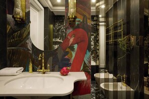 Een moderne badkamer met een witte wastafel, gouden details, een grote spiegel en een kleurrijke muurschildering.
